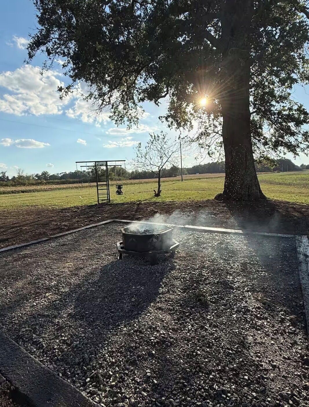 Un brasero rodeado de grava, un columpio y un árbol con luz solar filtrándose entre sus ramas.