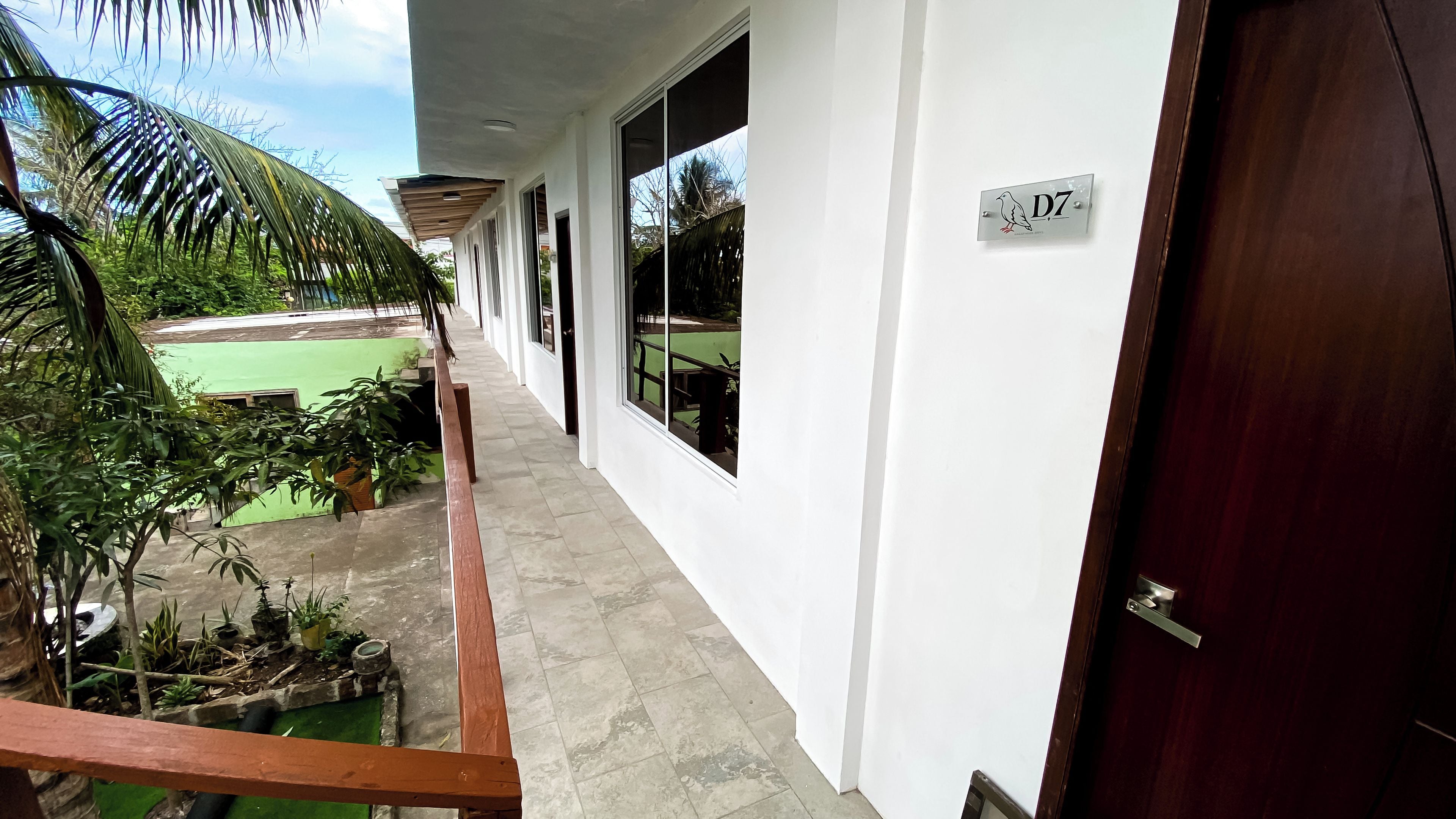 A corridor with a wooden railing, a door labeled 'D7', and a view of a green outdoor area with plants.