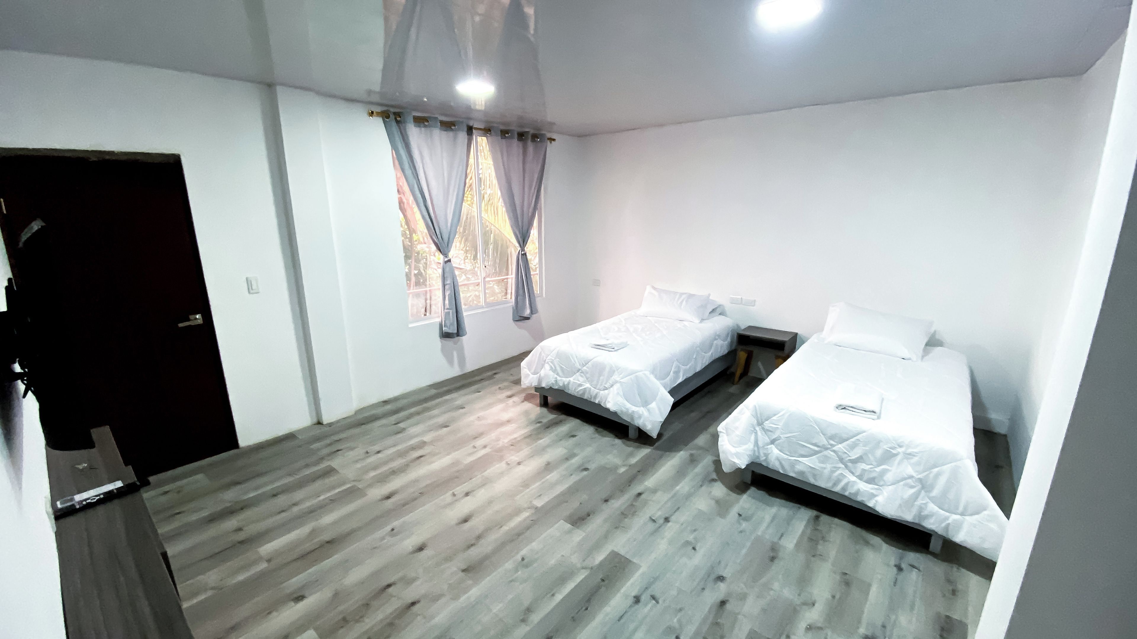 A room with two beds, a wooden floor, a window with curtains, and a door.