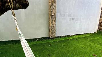 A hammock tied to a tree branch, a white wall, and a green artificial lawn.