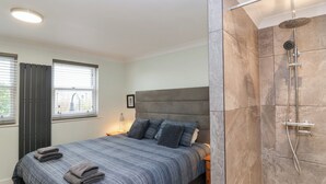 Ein modernes Schlafzimmer mit einem großen Bett, einem Nachttisch, einer Gardinenanlage am Fenster und einem Duschbereich mit Glastür.