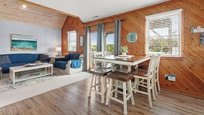 Een gezellige woonkamer met een blauwe bank, een houten eettafel met witte stoelen en uitzicht op het omliggende landschap door grote ramen.