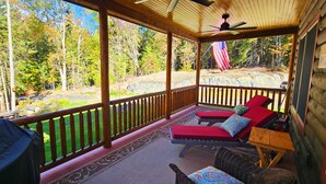 A covered porch with two red lounge chairs, a grill, and a view of a forested area.