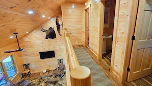 A wooden cabin interior with a loft bedroom, a ceiling fan, a mounted TV, and a rustic decor.
