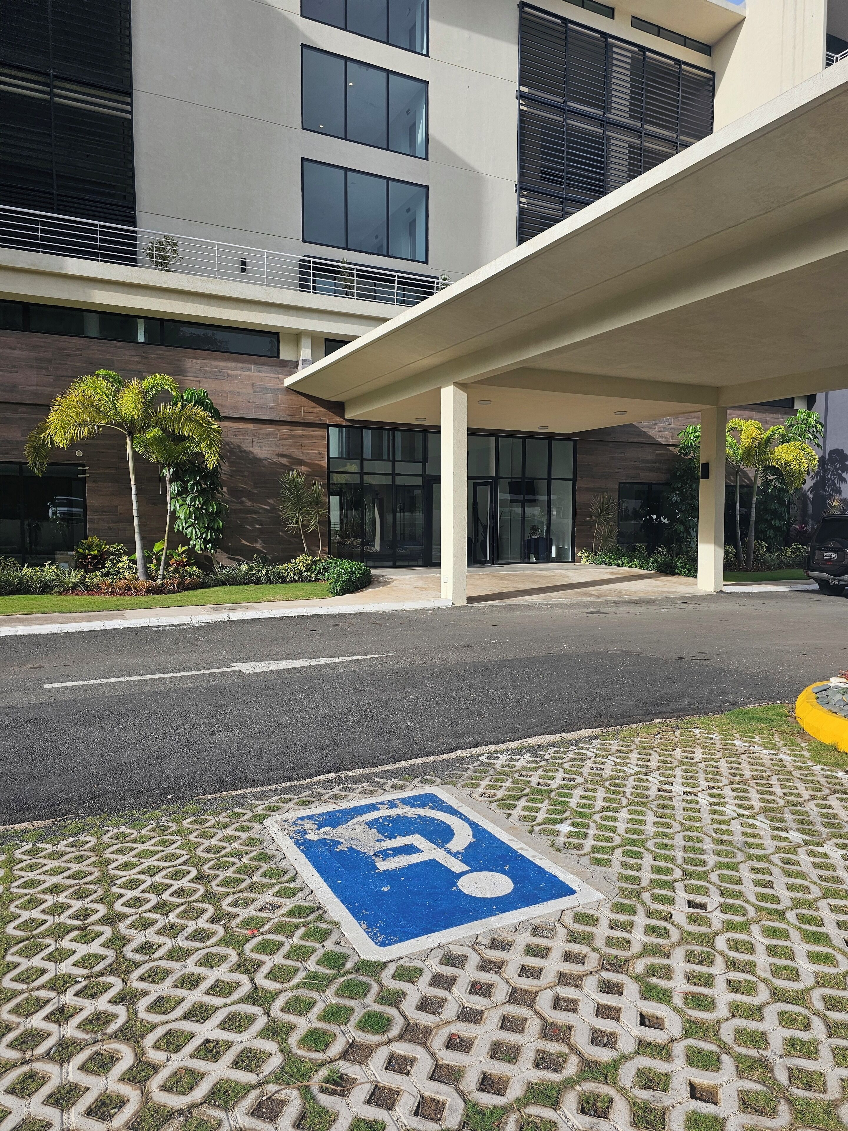 A modern building entrance with a covered walkway, a parking space marked with a wheelchair symbol, and a well-maintained lawn with palm trees.