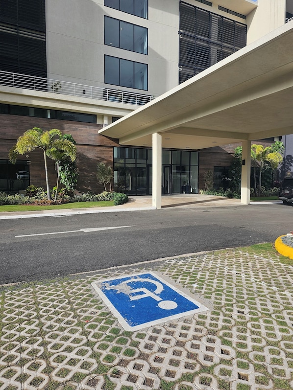 A modern building entrance with a covered walkway, a parking space marked with a wheelchair symbol, and a well-maintained lawn with palm trees.