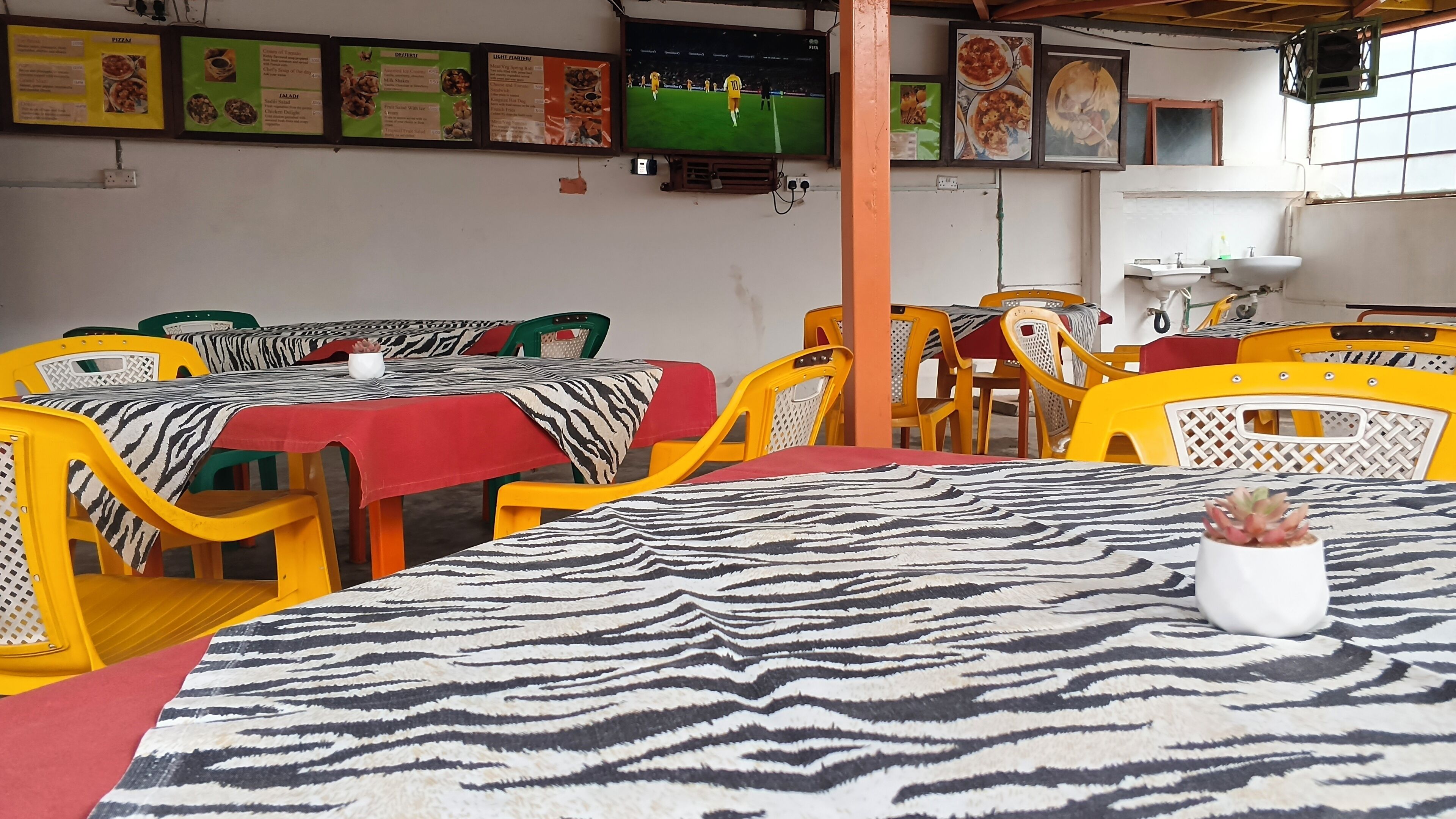 A restaurant with multiple tables and chairs, a television displaying a soccer match, and a menu board above the counter.