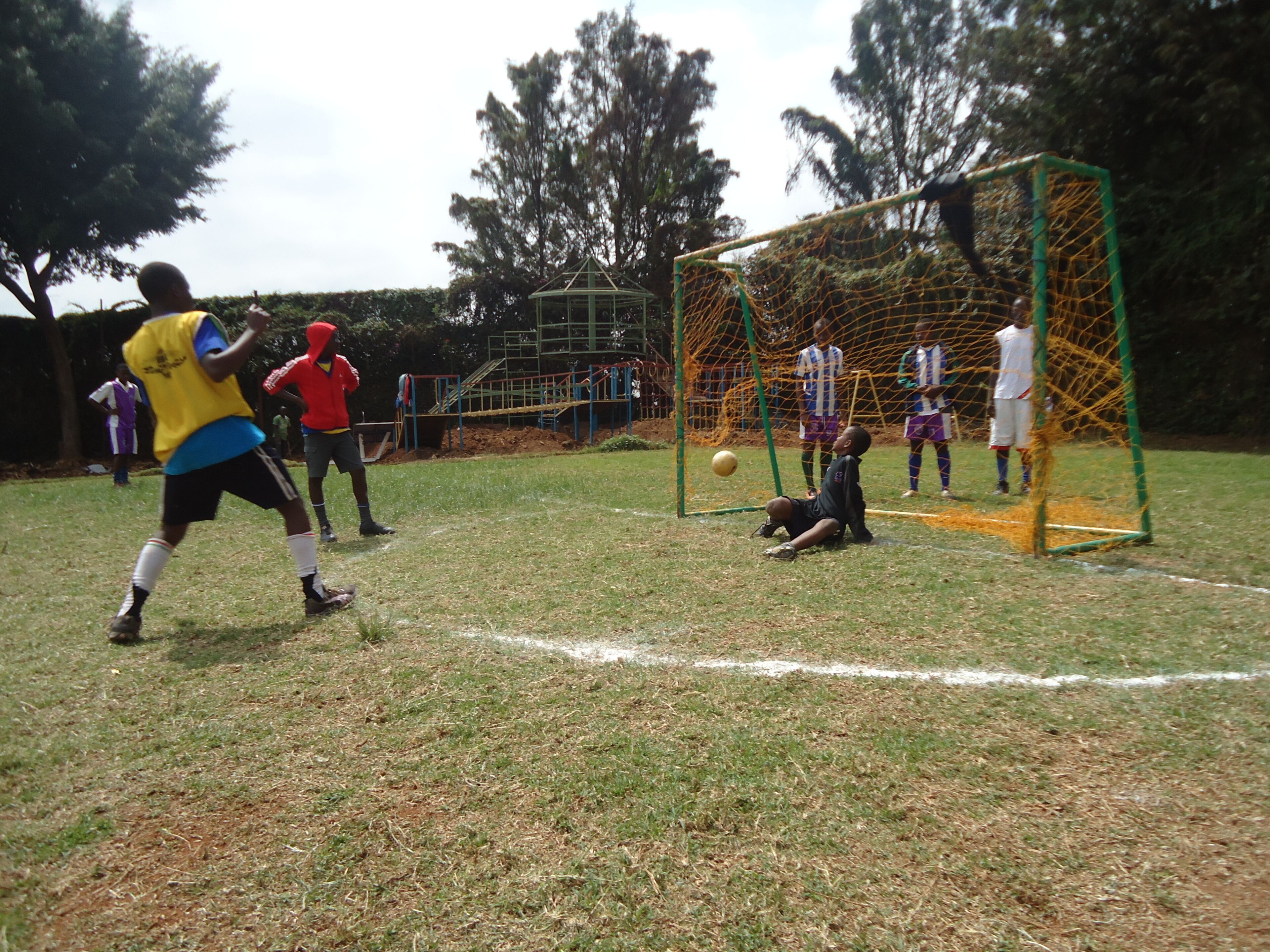 Partido de fútbol en desarrollo, con jugadores, un arco y un portero en un campo de césped.