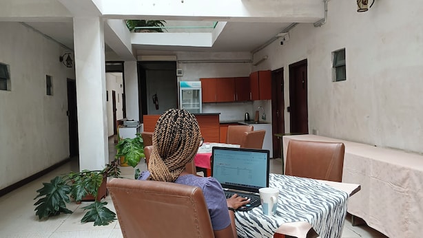 A person working at a laptop in a room with a dining table, chairs, and a kitchen area in the background. - Sadili Oval B & B (Nairobi)