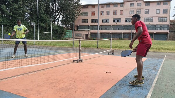 Una cancha de tenis con una red, dos jugadores y un edificio al fondo.