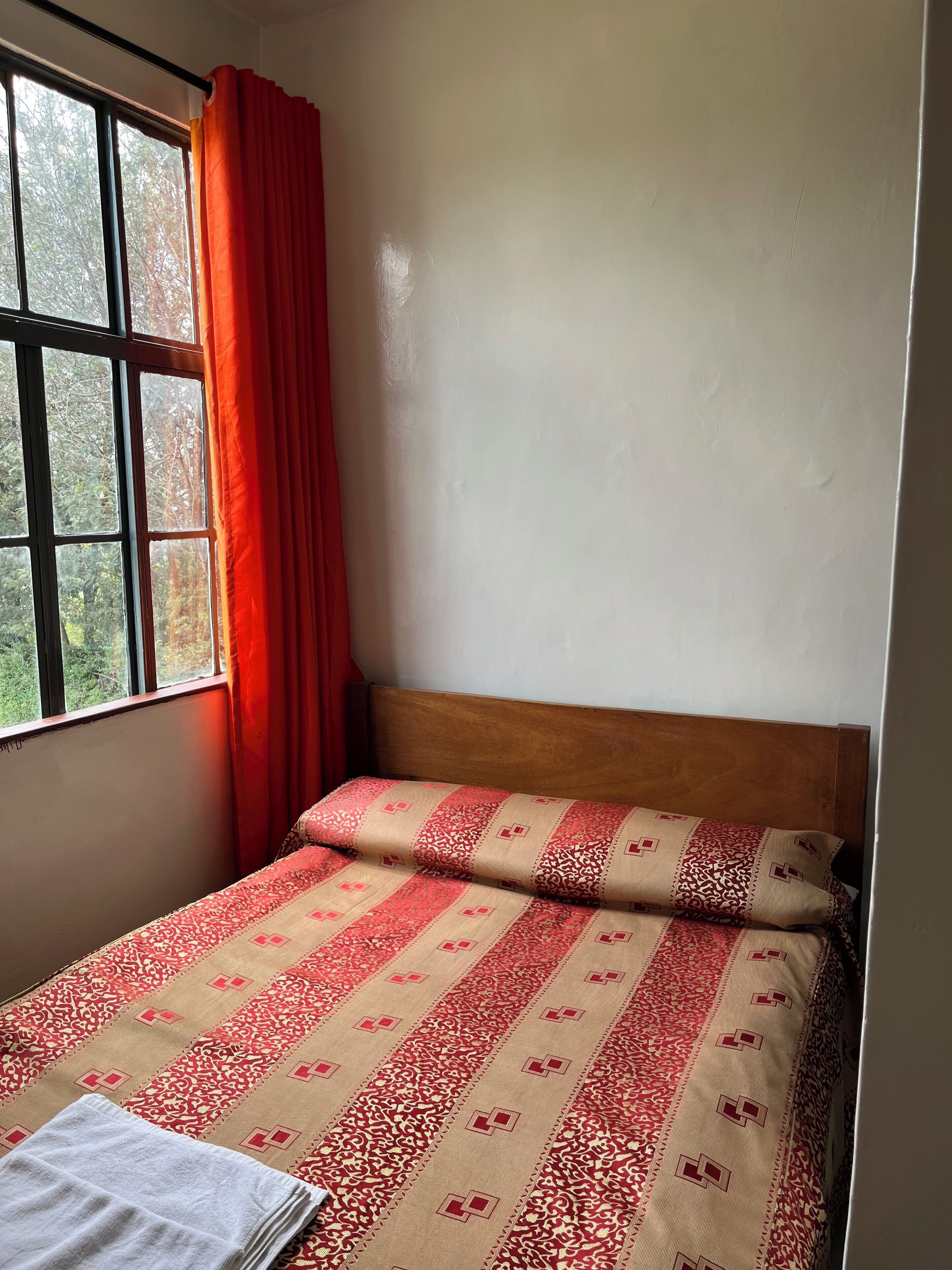 Una cama individual con una colcha estampada y almohadas, una ventana con cortinas, y una pared lisa.