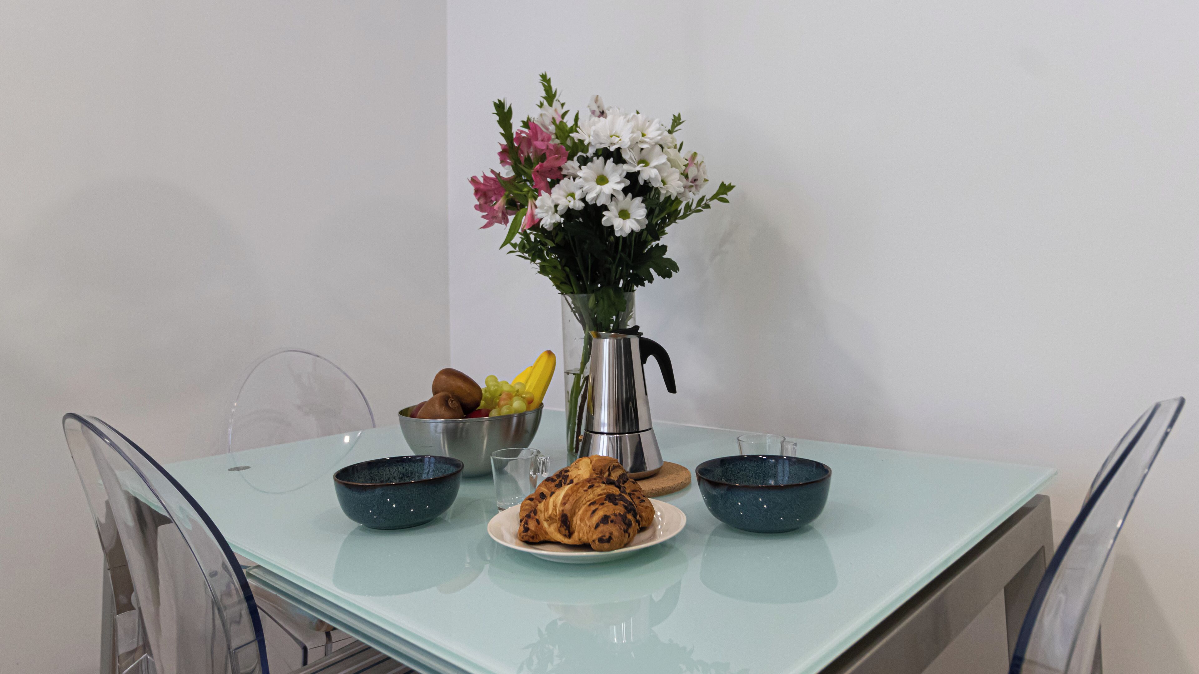 A dining table with a clear glass top, a metal pitcher, a bowl of fruit, a plate with croissants, and a vase with flowers.