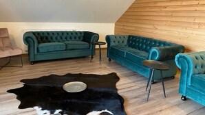 Ein Zimmer mit schräg gedämmter Decke und Holzverkleidung, ausgestattet mit zwei türkisfarbenen Sofas, einem schwarzen Lederteppich und einem kleinen runden Tisch.