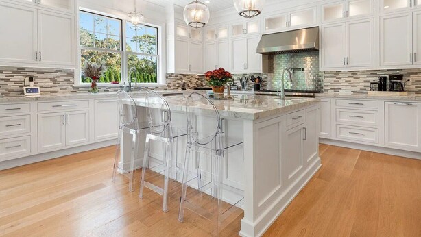 A spacious kitchen with white cabinetry, a large island with bar stools, a stainless steel range hood, and a view of the outdoors through a large window. - 6,400 square foot traditional-style home with a private pool and sweeping... (Southampton)