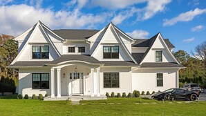 A large, two-story house with a symmetrical facade, a central entrance with a covered porch, and a driveway with two cars parked. - 6,400 square foot traditional-style home with a private pool and sweeping... (Southampton)