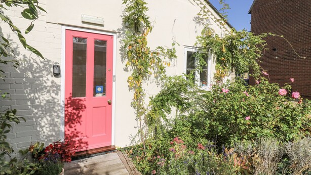 A white building with a red door and a small window. There is a brick building to the right and a garden with various plants and flowers. - Over Village Studio (Cambridge)
