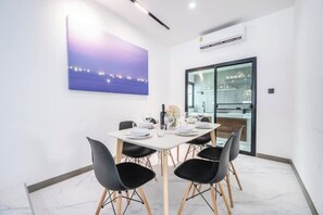 Un espace de salle à manger moderne avec une table blanche dressée pour quatre personnes, des chaises noires et un tableau représentant un paysage urbain accroché au mur.