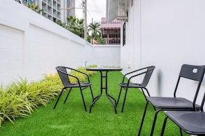 Un petit espace extérieur aménagé avec une table ronde et quatre chaises, entouré de gazon synthétique et de plantes.