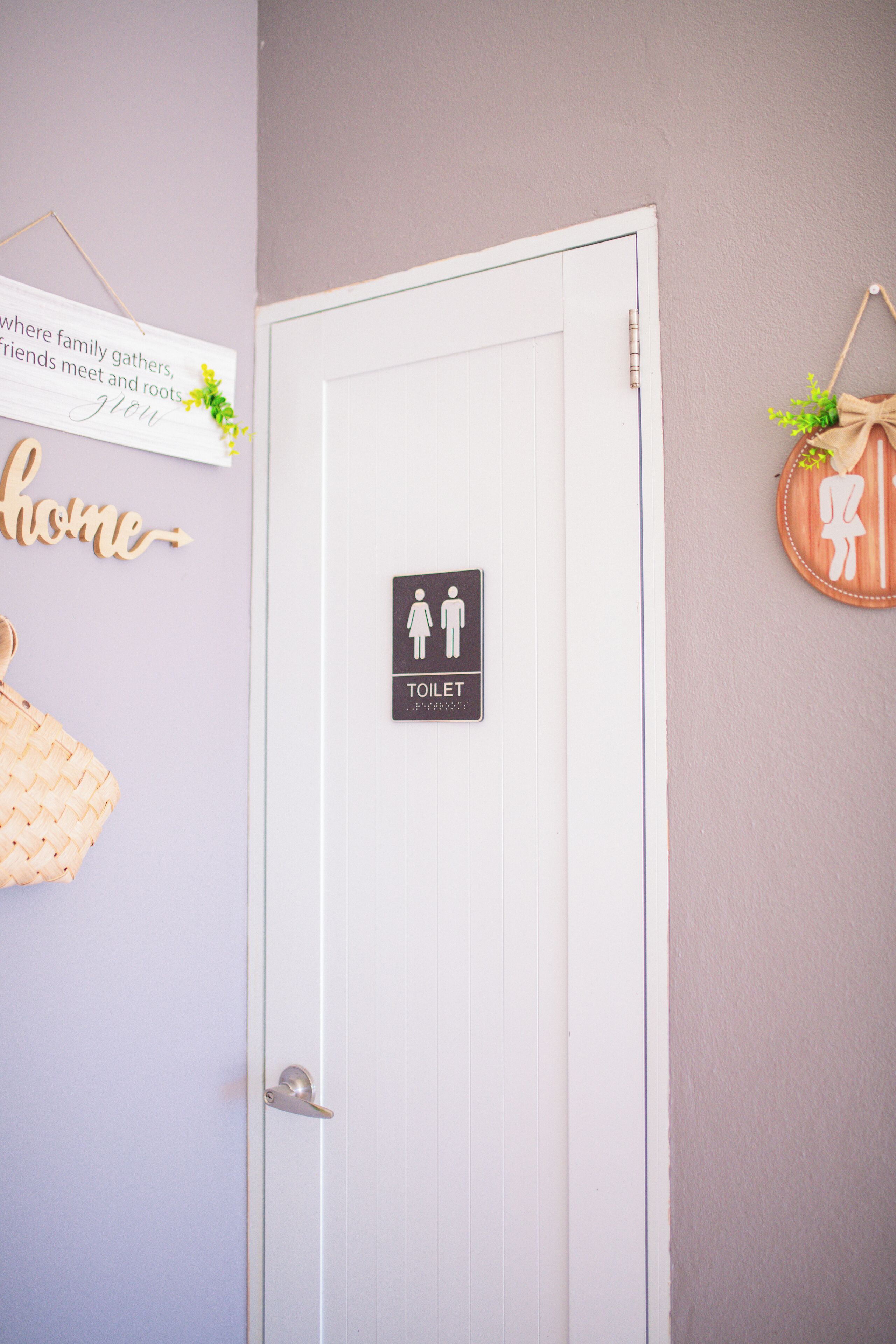 Een witte deur met een toiletbordje, een bord met de tekst 'waar familie samenkomt, vrienden elkaar ontmoeten en wortels groeien', en een wandmand aan de muur.