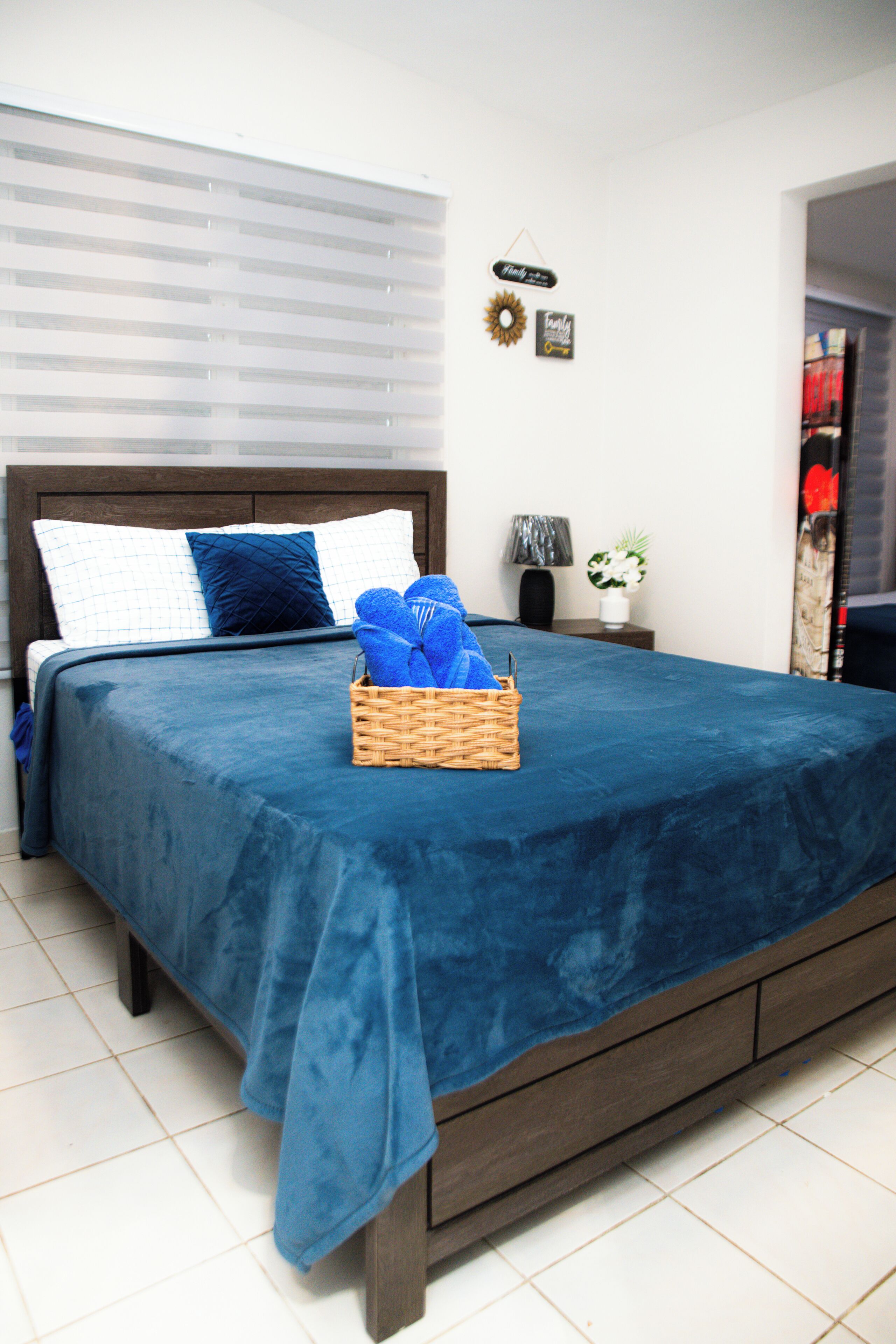 A neatly made bed with a blue bedspread, a wicker basket with towels, and a small bedside table with a lamp and a vase of flowers.