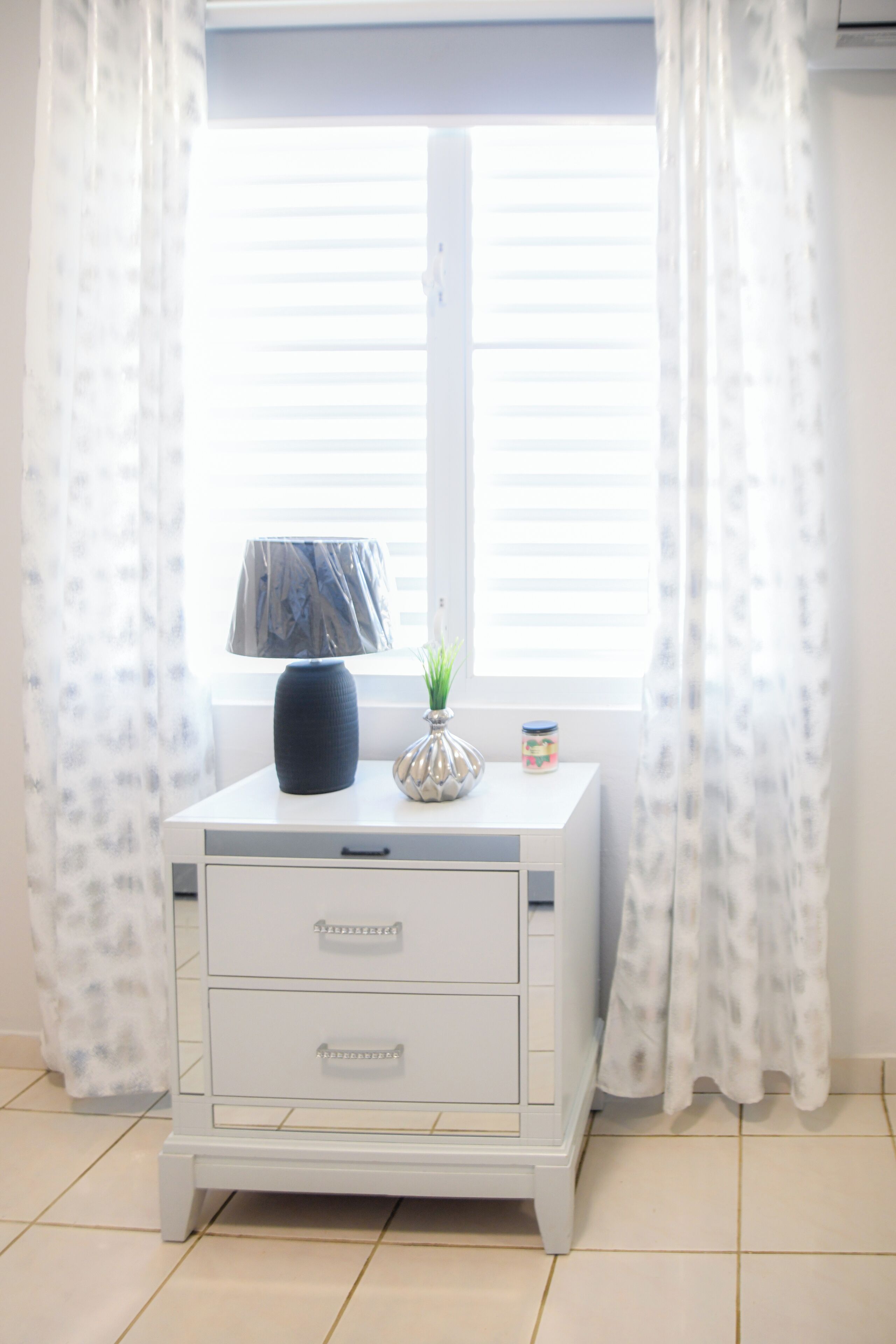 A white bedside table with drawers and a lamp, positioned by a window with white curtains.