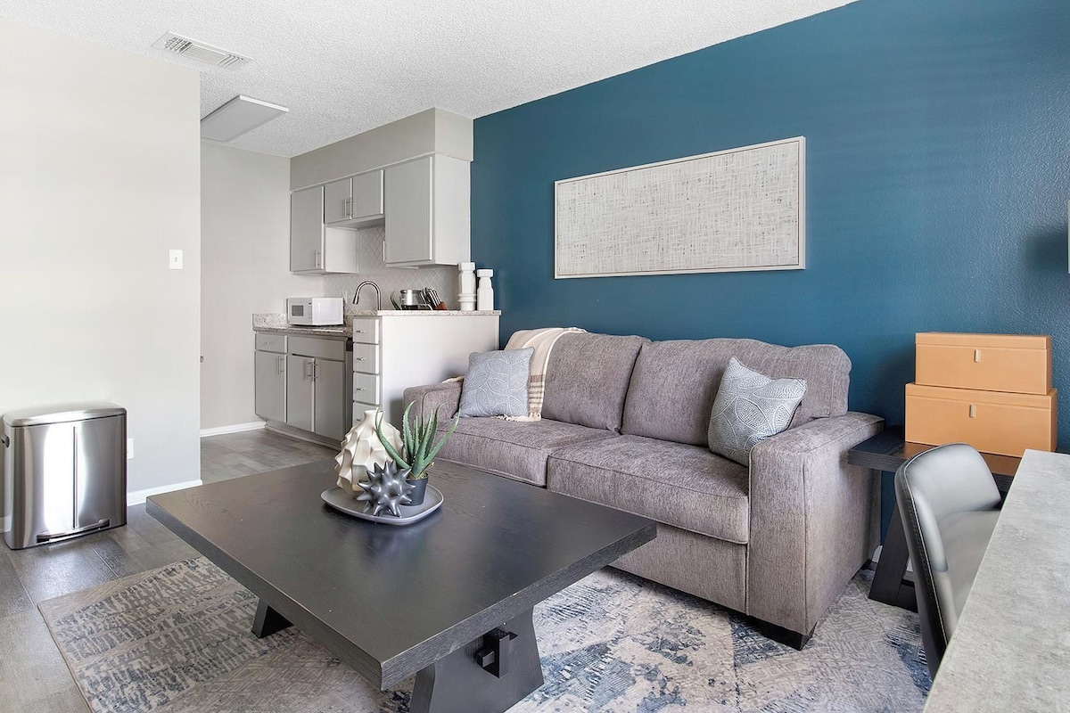 A modern living room with a grey sofa, a coffee table, and a kitchen area in the background.