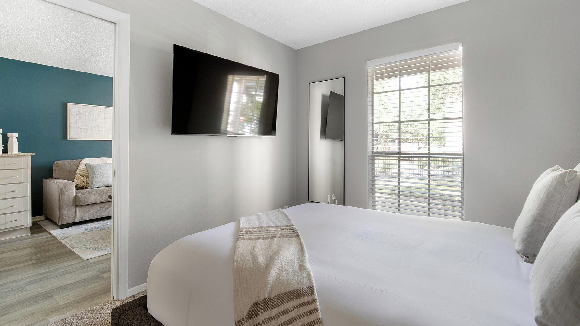 Een slaapkamer met een bed, een tv aan de muur, een dressoir en een raam met zonwering.