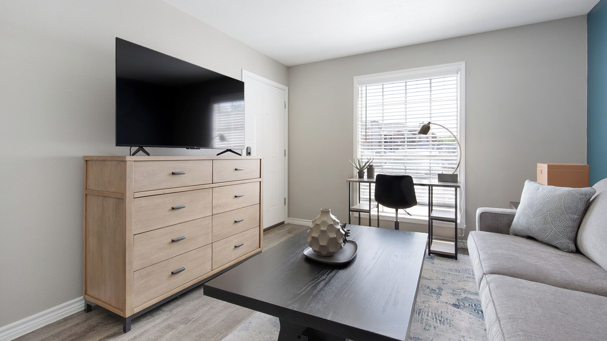 A modern living room with a flat-screen TV, a wooden cabinet, a coffee table, a sofa, and a window with blinds.