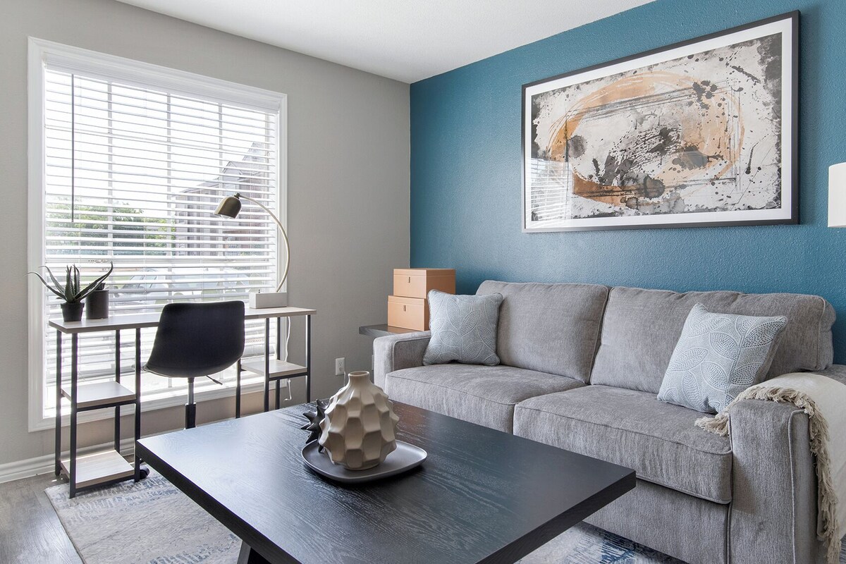 A living room with a grey sofa, a coffee table, a desk with a chair, a lamp, and a painting on the wall.