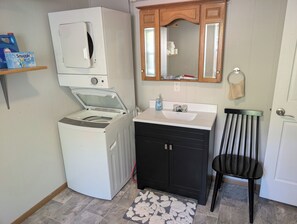 A compact bathroom with a washing machine, a sink, a mirror, and a chair.