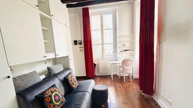 Una sala acogedora con un sofá gris oscuro, dos almohadones decorativos, un pequeño reposapiés y un escritorio blanco con una silla. Se ve una ventana con cortinas rojas y un radiador blanco.
