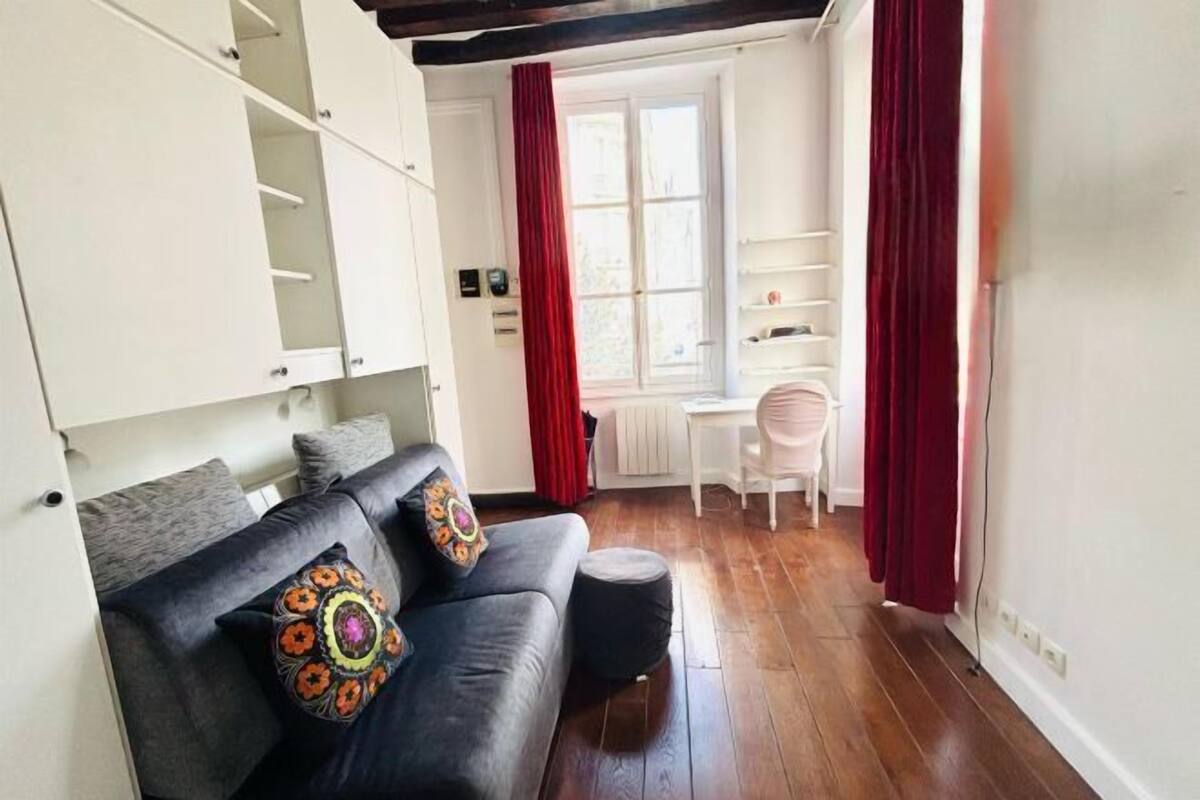 A cozy living room with a dark grey sofa, two decorative pillows, a small ottoman, and a white desk with a chair. A window with red curtains and a white radiator are visible.