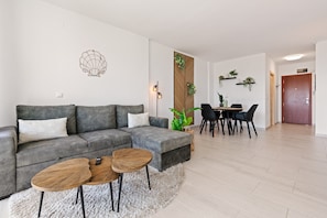 A modern living room with a grey sofa, two wooden coffee tables, a dining area with a black table and chairs, and a wooden door in the background.