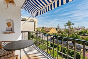 Un balcon avec une table et des chaises, un auvent à rayures et une vue sur une rue bordée de voitures garées et d’immeubles.