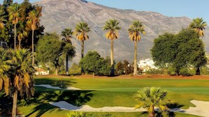 Un campo de golf con una montaña al fondo, palmeras y un edificio de paredes blancas.