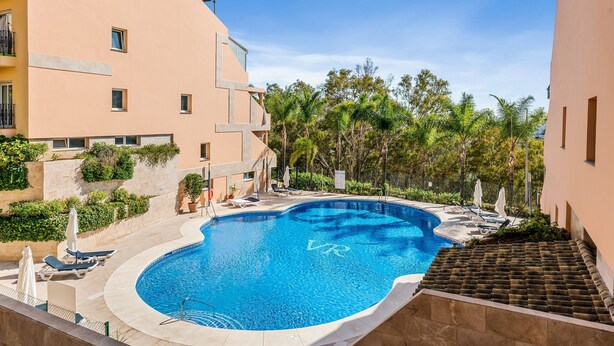 Una piscina rodeada de sillas de descanso y sombrillas, con un edificio al fondo.