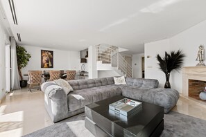 Un salon moderne avec un canapé d’angle gris, une table basse en verre, un escalier et une cheminée.