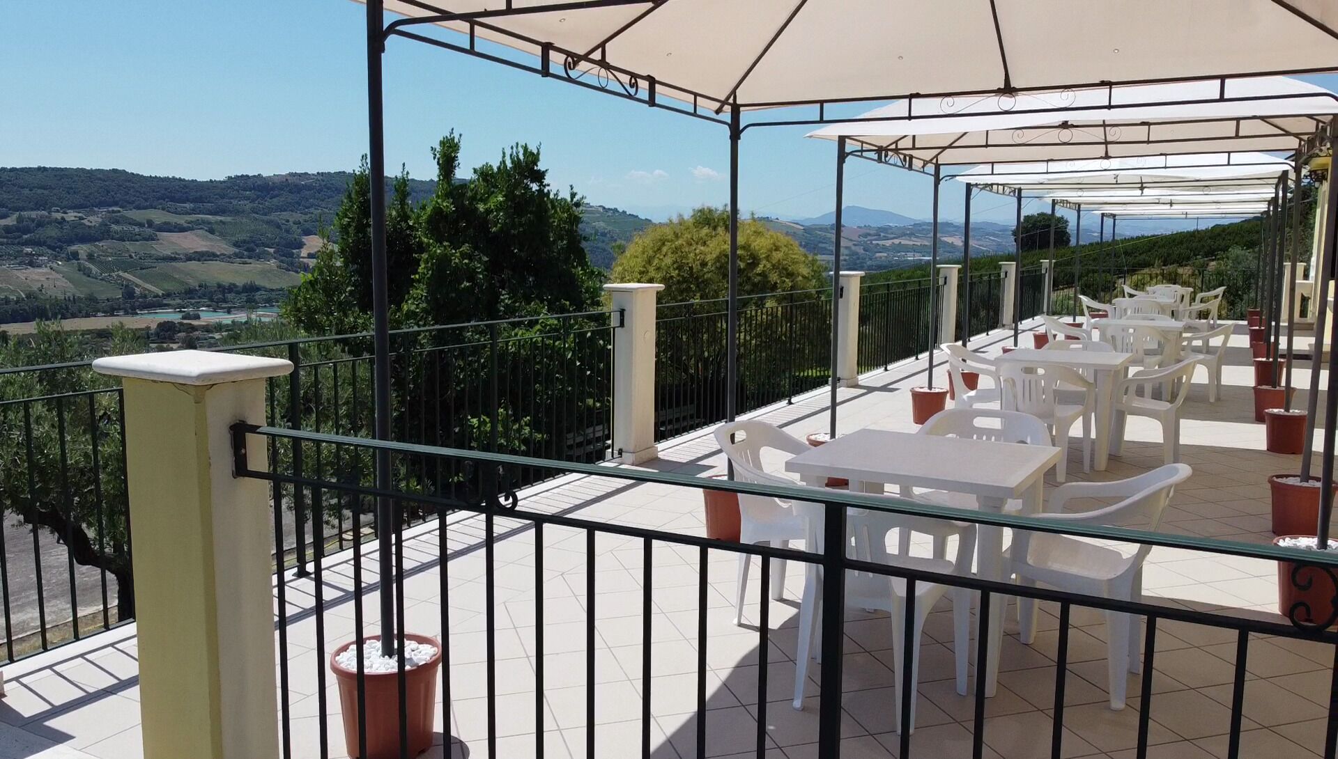 Una terraza con sillas y mesas de plástico blanco, una pérgola con dosel blanco y vistas al campo.
