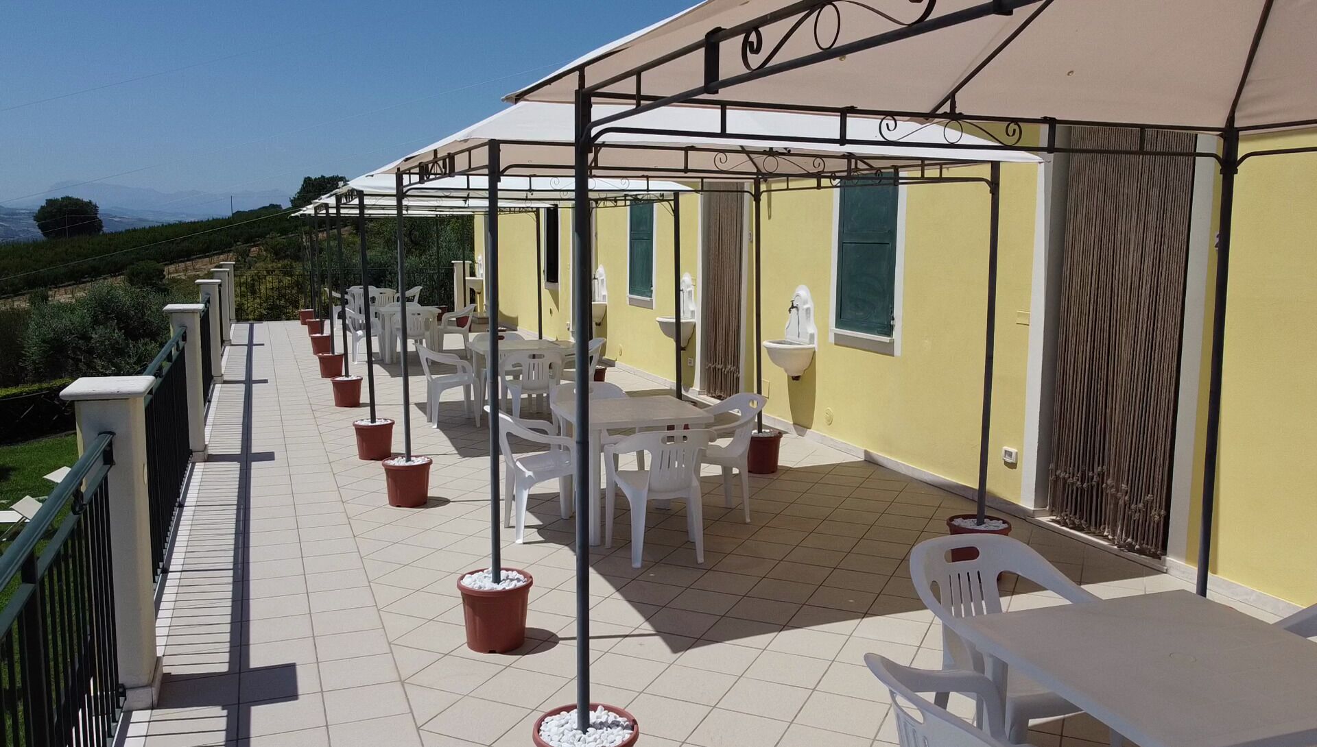 Un patio exterior con sillas y mesas de plástico blanco, una zona cubierta con un toldo color beige y una pared amarilla con ventana y lavamanos.