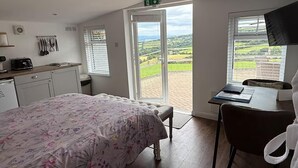 A bedroom with a bed, a desk, and a view of a green landscape through a sliding door. - 1 Bed Cabin-sleeps 2, Hot Tub, Garden, Pets, Views (Callington)