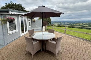 A patio with a round table and wicker chairs, a dark umbrella, and a view of green fields. - 1 Bed Cabin-sleeps 2, Hot Tub, Garden, Pets, Views (Callington)