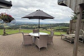 A patio with wicker furniture and a table under a large umbrella, overlooking a scenic countryside view. - 1 Bed Cabin-sleeps 2, Hot Tub, Garden, Pets, Views (Callington)