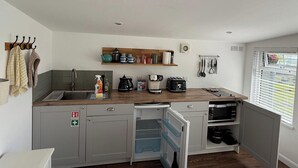 A compact kitchen with a sink, a coffee maker, a toaster, and a microwave. There is a window with blinds, a shelf with kitchen utensils, and a first aid sign on the cabinet. - 1 Bed Cabin-sleeps 2, Hot Tub, Garden, Pets, Views (Callington)