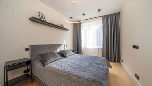 A modern bedroom with a large bed, a bedside table, a built-in shelf with decorative items, and a large window with curtains. - Beach and forest front apartment in Dzintari (Jūrmala)