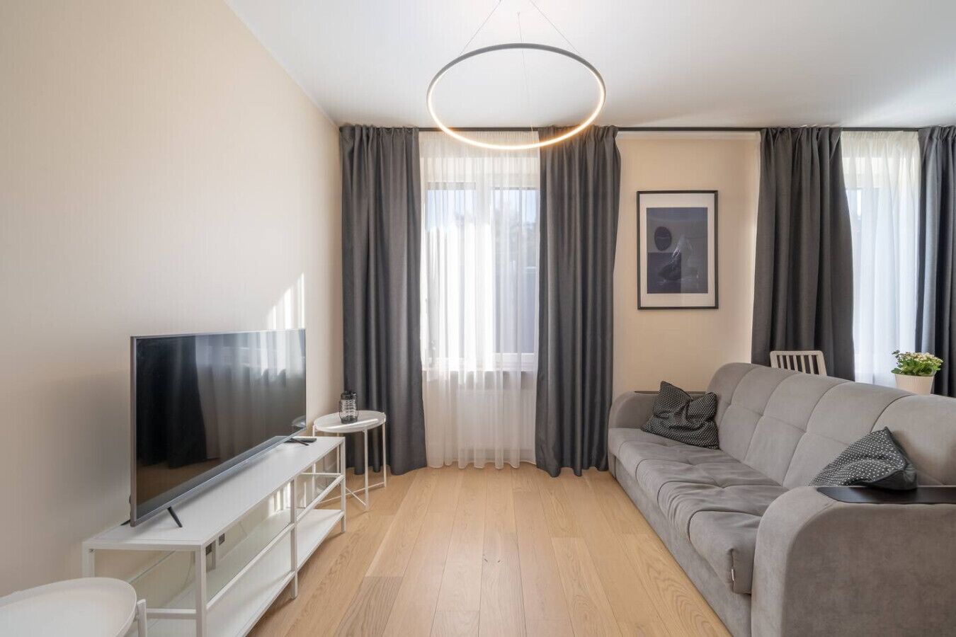 A modern living room with a flat-screen TV on a white stand, a grey sofa with cushions, a small round table with a lamp, and a large window with grey curtains.