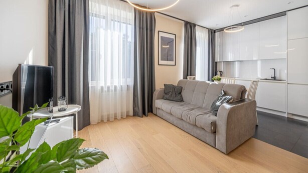 A modern living room with a grey sofa, a small side table, and a large window with sheer curtains. - Beach and forest front apartment in Dzintari (Jūrmala)