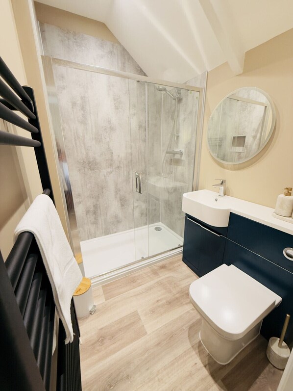 A modern bathroom with a glass shower enclosure, a white toilet, a sink with a dark cabinet, and a round mirror on the wall.