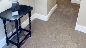 A hallway with beige walls, a dark wooden table with a lamp, and a carpeted floor. - Rising Sun Inn - Bronn Ewhella (Launceston)