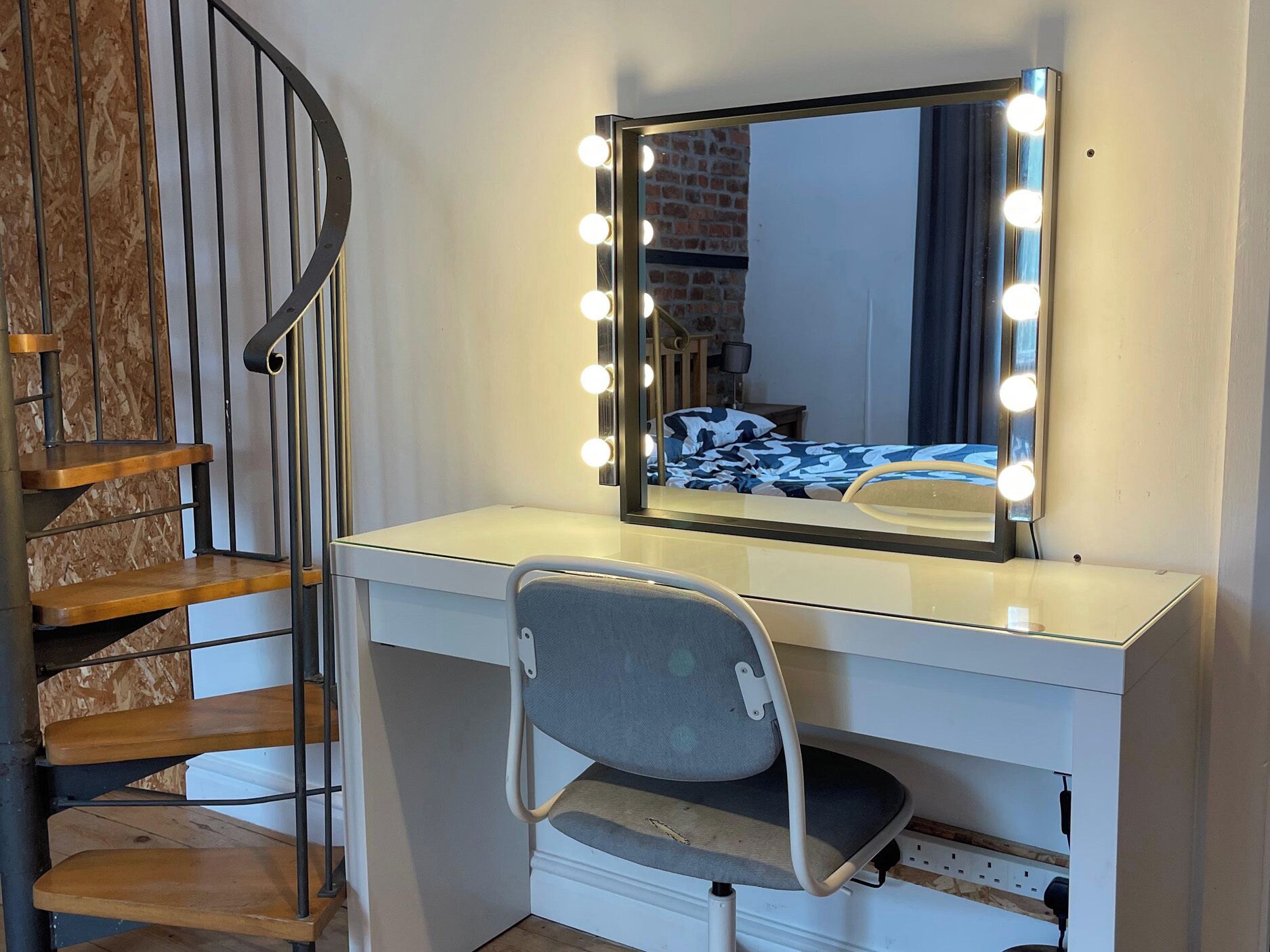 A room with a vanity, a chair, a mirror with lights, and a spiral staircase.