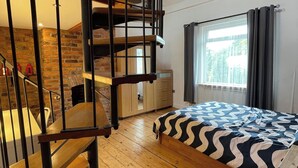 Ein Schlafzimmer mit einem hölzernen Bett, einem Fenster mit Vorhängen, einer Ziegelwand und einer Treppe mit Geländer.